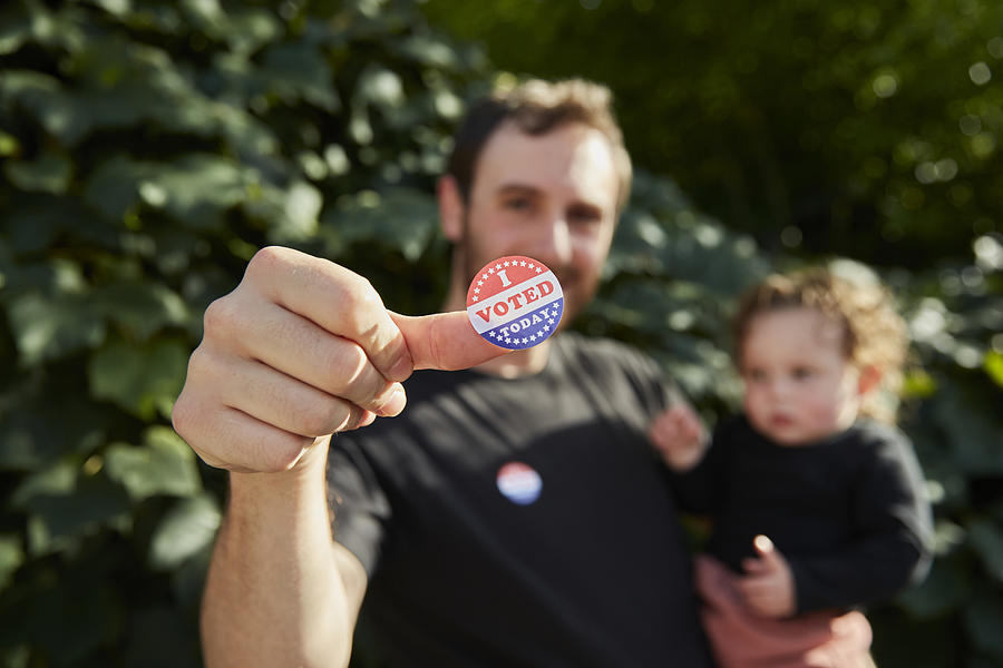A father standing outside holding his baby, with an I Voted Sticker on his thumb Photograph by EyeWolf
