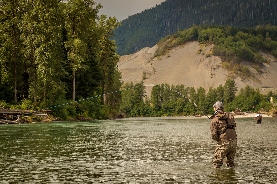 A fly fisherman hooked into a fish on a river with mountains and trees ...