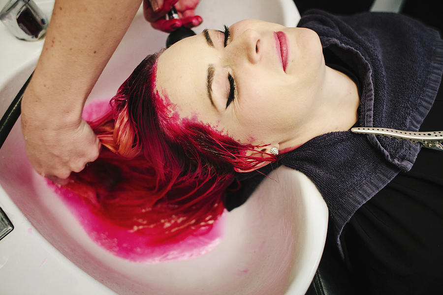A hair salon client having red hair dye rinsed from her hair over a basin. Photograph by Mint Images