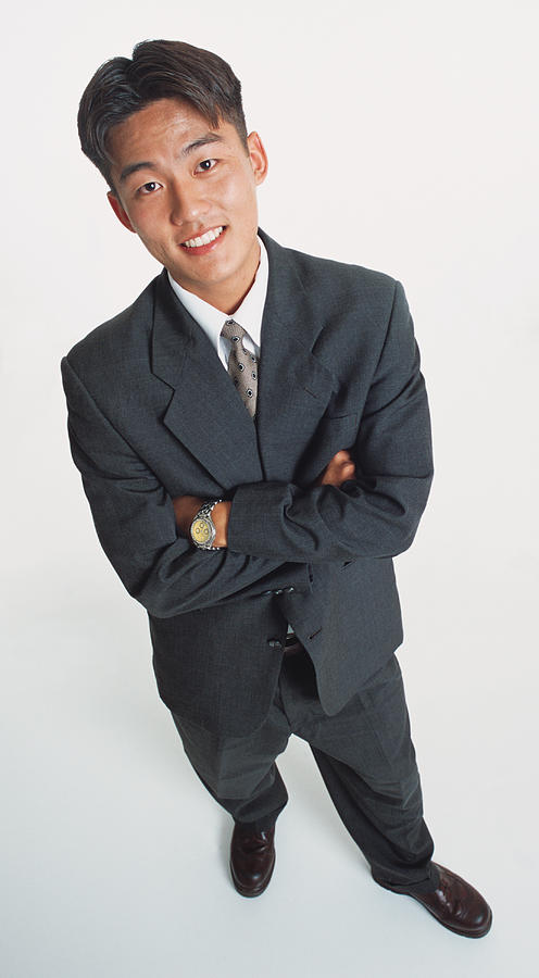 A Handsome Young Asian Man Wearing A Dark Suit And Standing With Arms Crossed Looks Up At The Camera Photograph by Photodisc