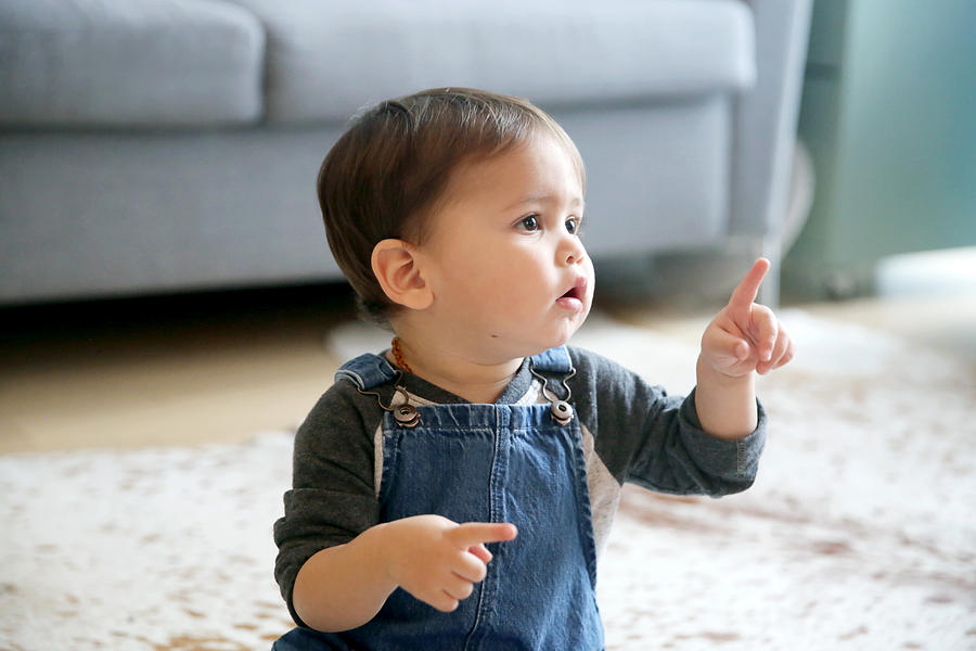 A LatinX toddler points his index finger, while sitting in his living room wearing jean overalls and a long sleeved t-shirt. Photograph by Mireya Acierto