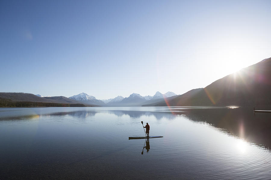 A man stand up paddle boards (SUP) on a calm Lake McDonald in Glacier National Park. Photograph by Craig Moore