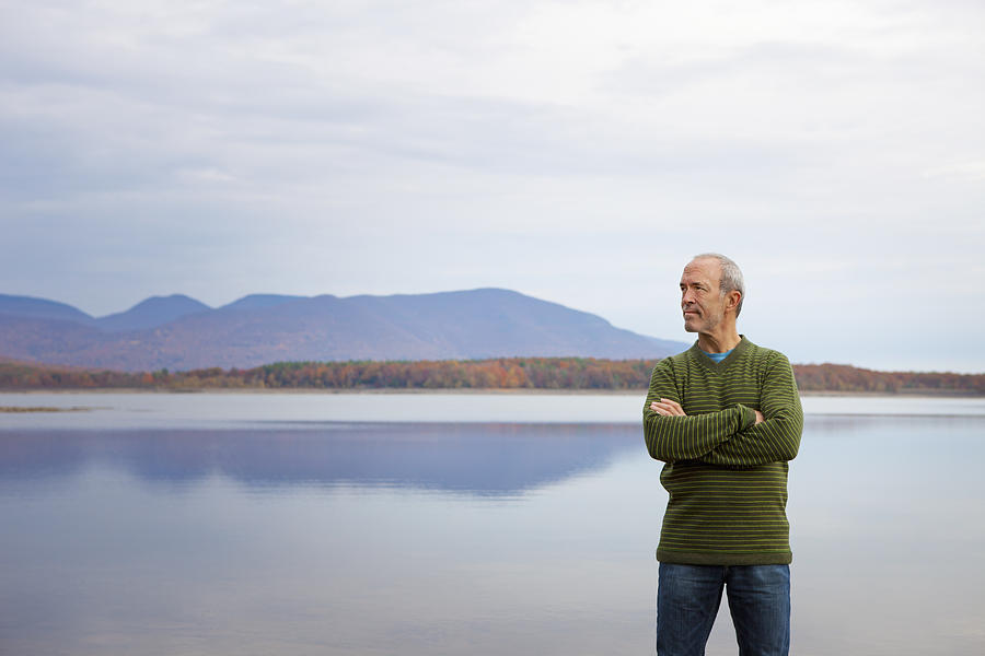 A man with arms folded by the water of a flat calm lake in the mountains. Photograph by Mint Images - Bill Miles