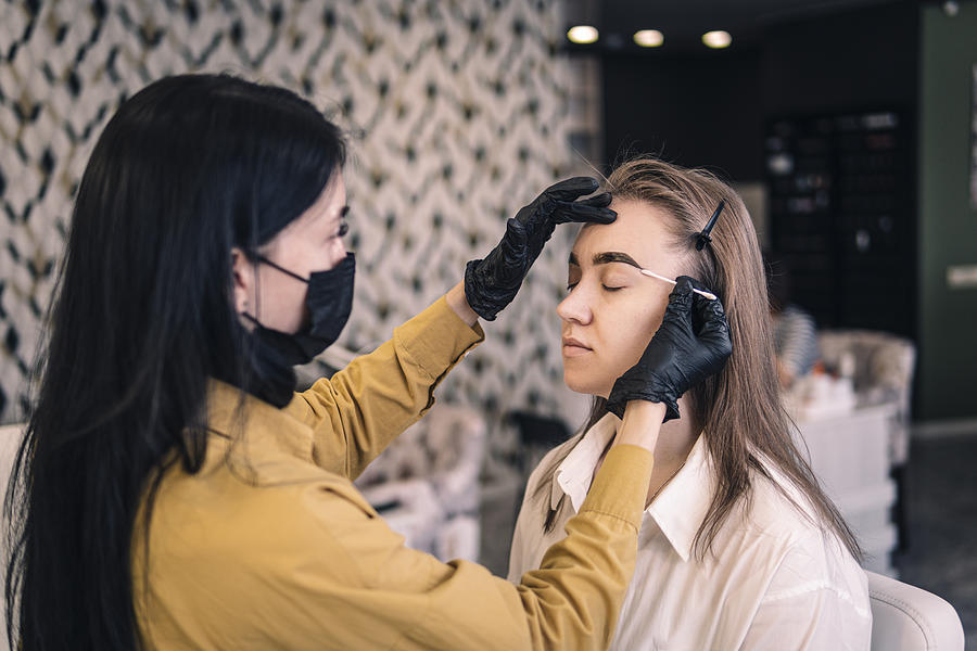 A master cosmetologist paints the eyebrows of a client in the salon close-up. eyebrow dyeing procedure, girl wipes paint from her eyebrows. Photograph by Diy13