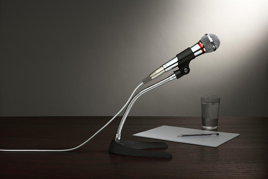 A microphone in a stand, with paper and a glass of water, all on a wooden desktop Photograph by Creative Crop