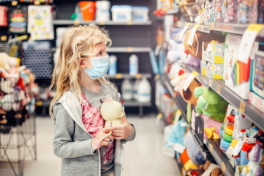 A new normal. Caucasian blonde girl in sanitary face mask shopping at toy store. Child wearing protective mask against coronavirus. Safety, health protection during covid-19 quarantine. Photograph by ~UserGI15613517