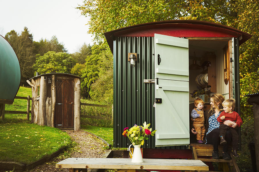 A shepherds hut with open door beside a path to a small rustic shed, and a woman with two small children seated on the step. Photograph by Mint Images