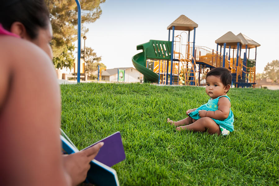 A woman holding a childrens book tries to engage the attention of a baby girl sitting in the lawn of a kids playground. Photograph by Mmg1design