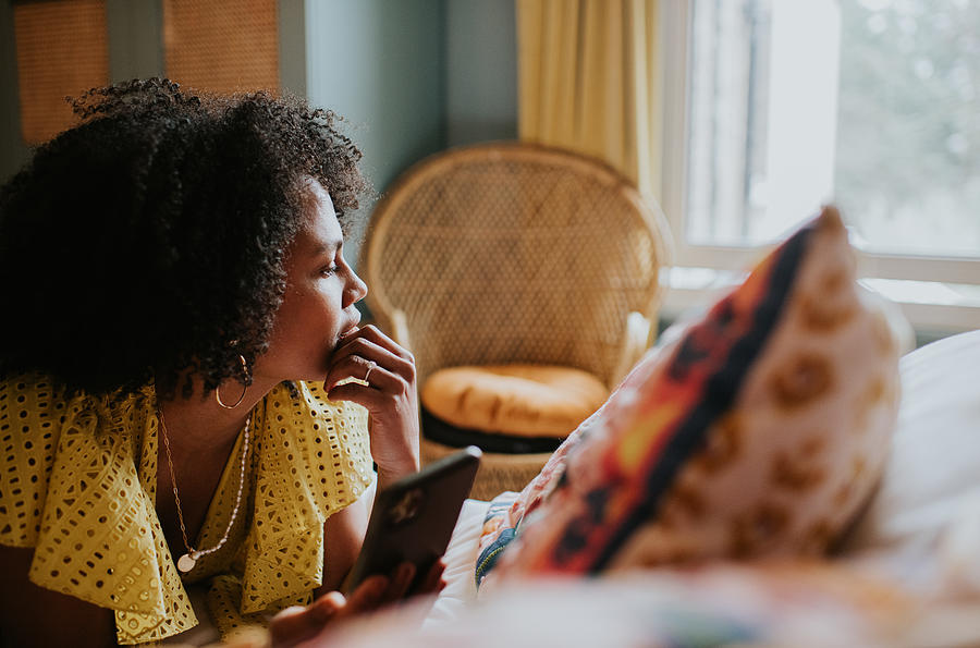 A woman looks distracted and pensive as she lies on a bed holding a mobile phone, and gazes out the window Photograph by Catherine Falls Commercial