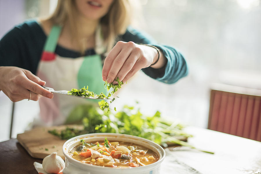 A woman wearing an apron, sitting at a table, sprinkling herbs into a bowl of vegetable stew. Photograph by Mint Images