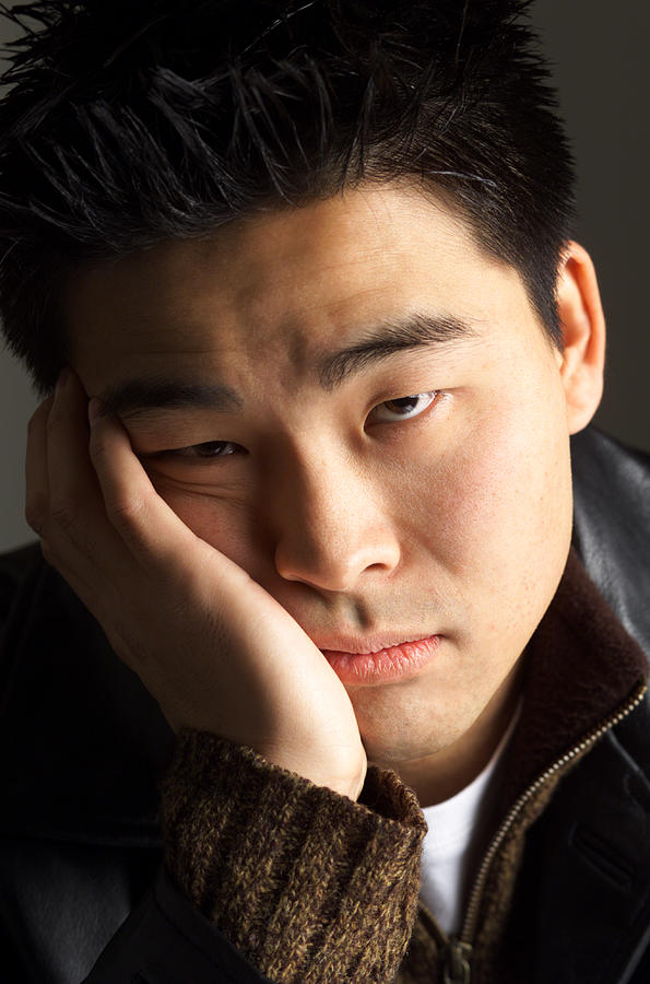A Young Asian Man With Spiked Hair And A Leather Jacket Is Leaning His Cheek In His Hand And Looking Discouraged Photograph by Photodisc