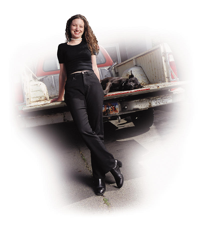 A Young Caucasian Woman In Black Pants And Shirt Is Leaning Against The Tailgate Of A Pickup Truck With Her Black Dog Lying In The Truck Bed Photograph by Photodisc