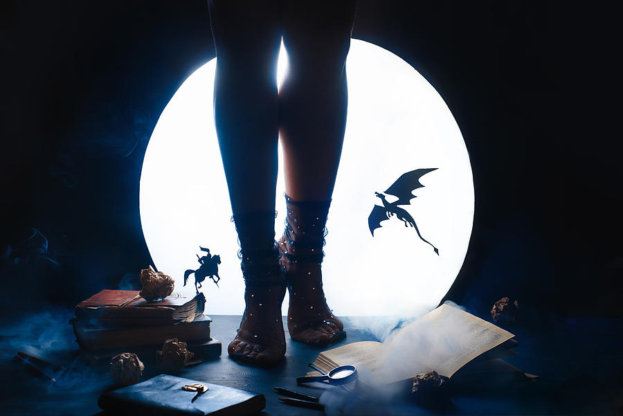 A young female writer with a fairy tale book. Dragon and Knight silhouettes against shiny Moon background. Legs in starry socks Photograph by Dina Belenko Photography