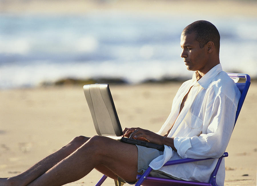 A Young Man In Tan Shorts And A White Oxford Shirt Is Reclining In A Beach Chair Typing In To A Laptop Computer Photograph by Photodisc