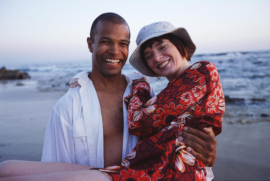 A Young Man Is Carrying A Young Woman In His Arms With The Beach And Shoreline Behind Them Photograph by Photodisc