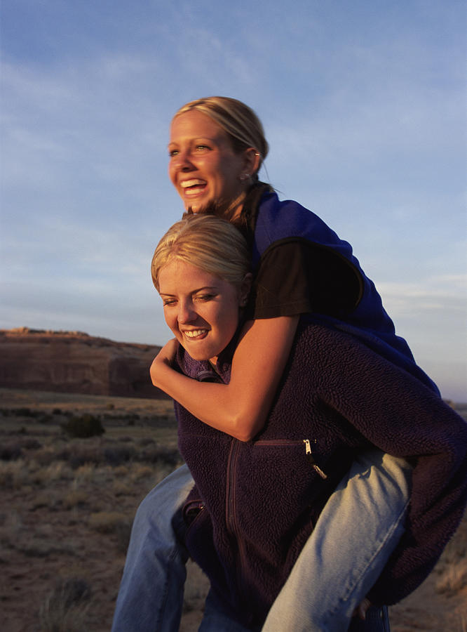 A Young Woman In Blue Jeans And A Purple Fleece Is Giving Her Friend Who Is Wearing Blue Jeand And A Blue Fleece Vest A Piggyback Ride With The Southern Utah Desert In The Background Photograph by Photodisc