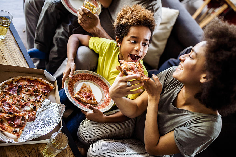 Above view of black mother feeding her daughter with pizza at home. Photograph by Skynesher Above view of black mother feeding her daughter with pizza at home. Photograph by Skynesher