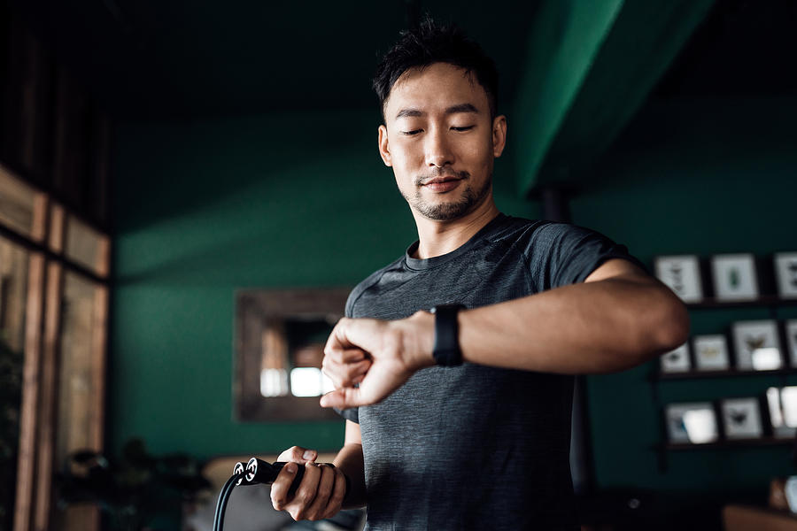 Active young Asian man exercising at home, using fitness tracker app on smartwatch to monitor training progress and measuring pulse. Keeping fit and staying healthy. Health, fitness and technology concept Photograph by AsiaVision