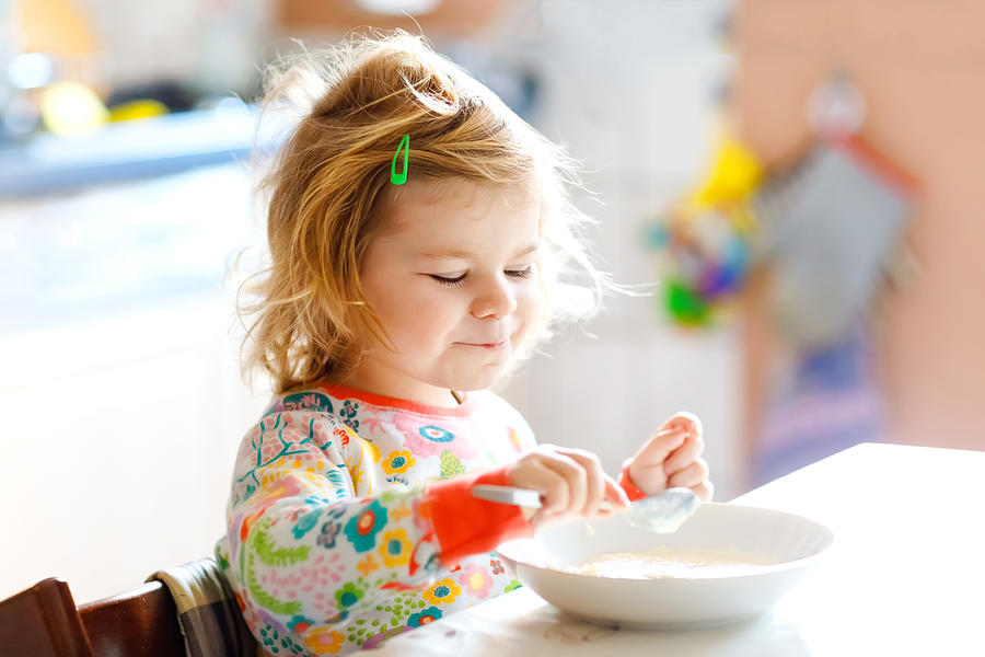 Adorable toddler girl eating healthy porridge from spoon for breakfast. Cute happy baby child in colorful pajamas sitting in kitchen and learning using spoon. Photograph by Romrodinka