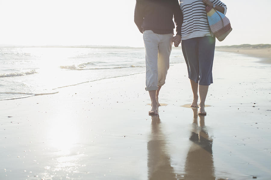 Affectionate barefoot mature couple walking, holding hands in sunny ocean beach surf Photograph by Caia Image