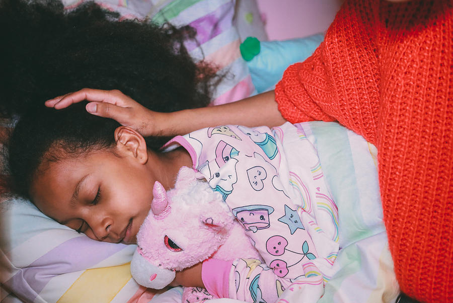 Afrolatina girl in pajamas holding stuffed unicorn with her eyes closed. Mothers face and neck not visible. Mothers hand on daughters head. Photograph by Evelyn Martinez