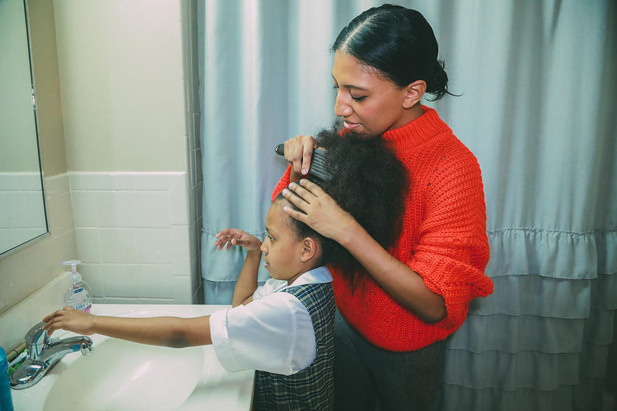 Afrolatina mom with Afrolatina daughter in the bathroom. Mom is combing daughters afro. Daughter reaches out for faucet handle. Photograph by Evelyn Martinez