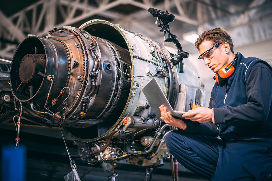 Aircraft engineer in a hangar using a laptop while repairing and maintaining an airplane jet engine Photograph by Extreme-photographer