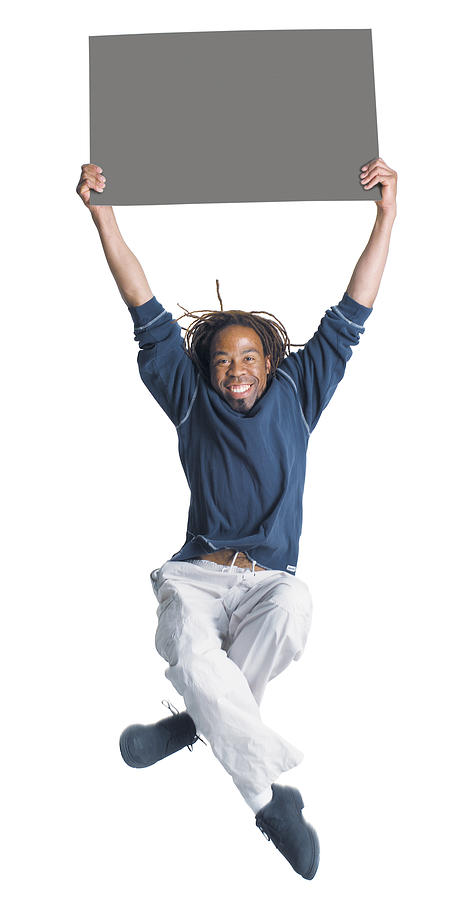 An African American Man With Dreadlocks Wearing Kakhi Pants And A Blue Shirt Jumps In The Air With Is Legs Crossing As He Holds A Blank Sign Above His Head With Two Hands Photograph by Photodisc