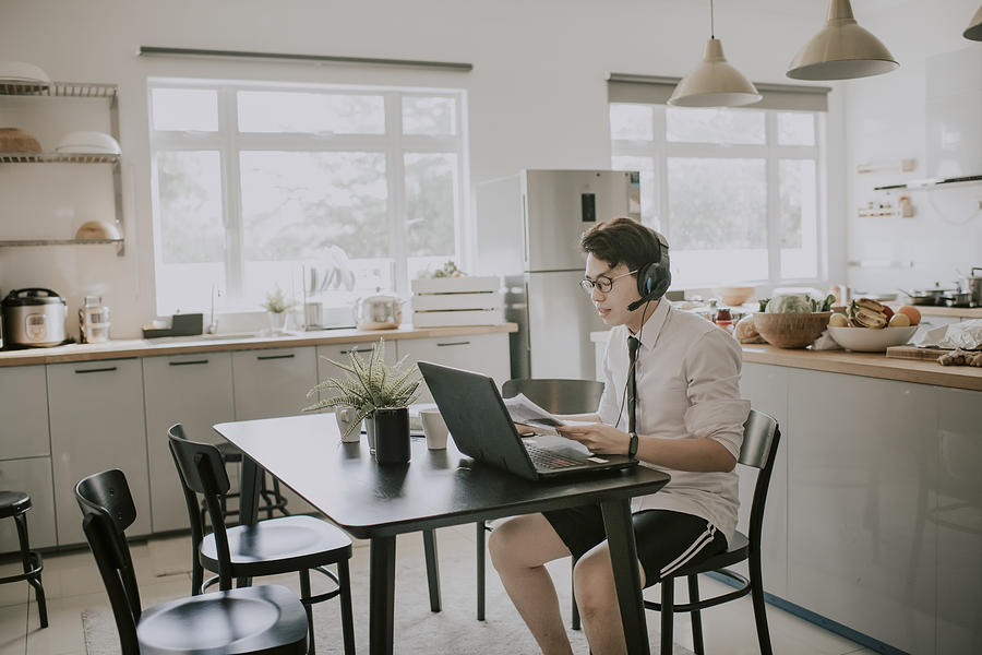 An asian chinese young adult man with tie, long sleeve shirt and short pant sitting using laptop in his dining room for video conference with his business partner and colleague virtual business meeting Photograph by Chee Gin Tan