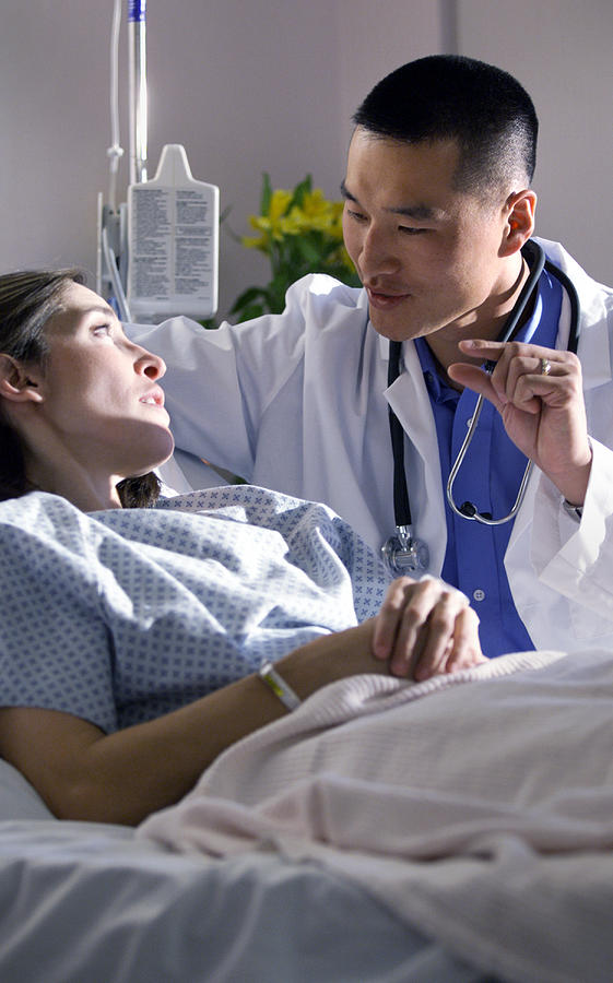 An Asian Doctor Advises A Caucasian Female As She Lies In A Bed In A Recovery Room Photograph by Photodisc