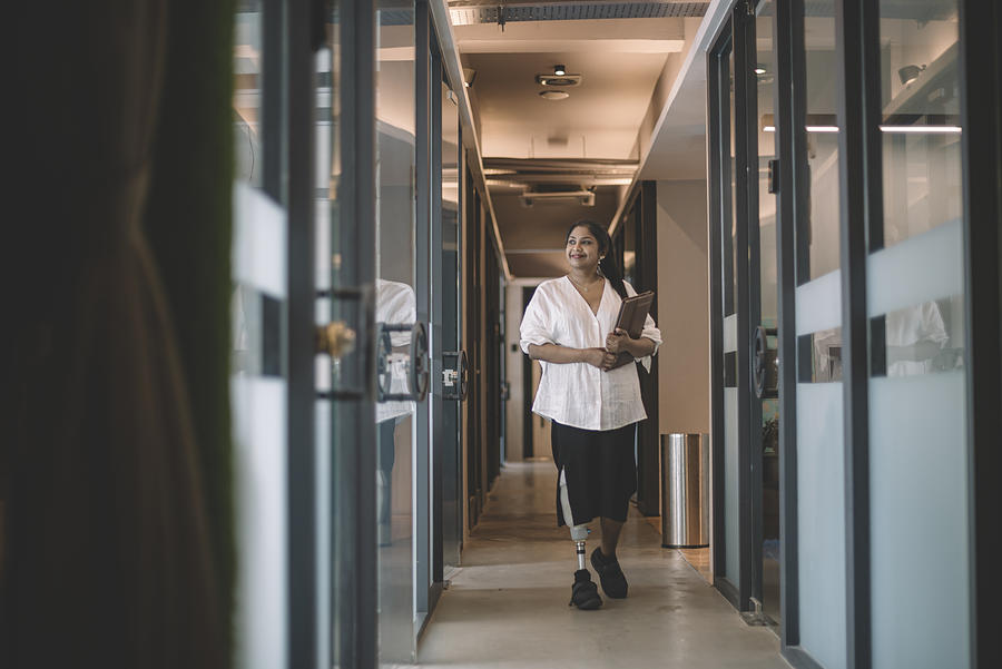 An Asian Indian Female Handicapped White Collar Worker With Prosthetic Limb  Holding Her Digital Tablet And Walking At The Office Corridor Smiling Photograph by Edwin Tan
