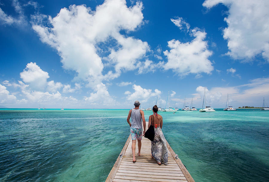 ANEGADA ISLAND, BRITISH VIRGIN ISLANDS, CARIBBEAN. A couple walks down a long dock surrounded by teal sea and blue sky. Photograph by David Hanson