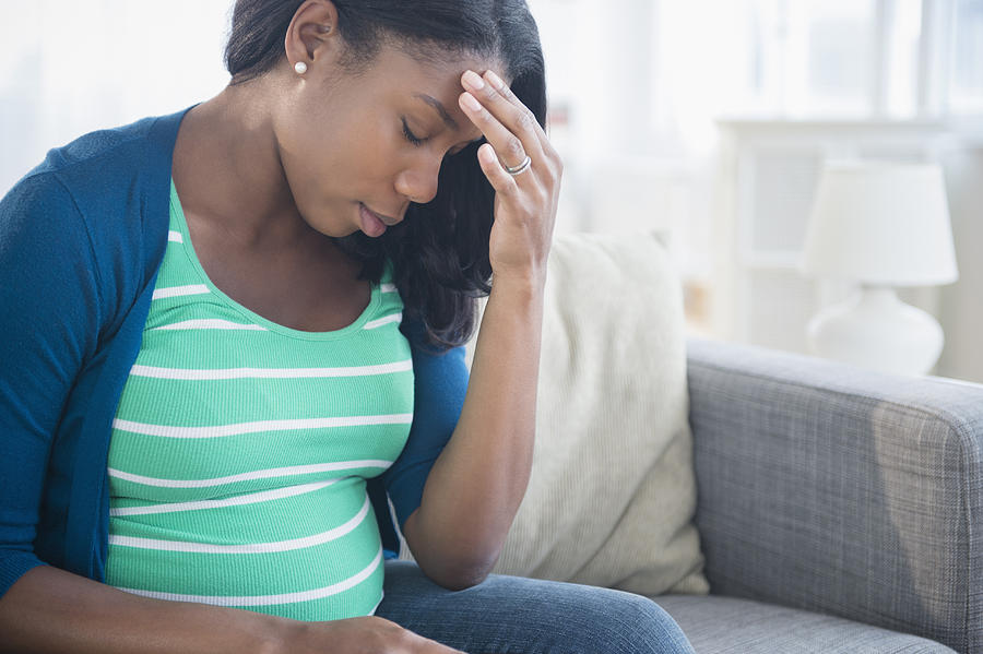Anxious Black pregnant woman rubbing forehead on sofa Photograph by JGI/Jamie Grill