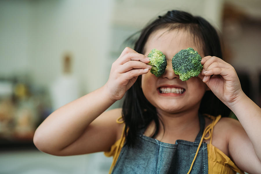 Asian chinese female child act cute with hand holding broccoli putting in front of her eyes with smiling face at kitchen Photograph by Marcus Chung 