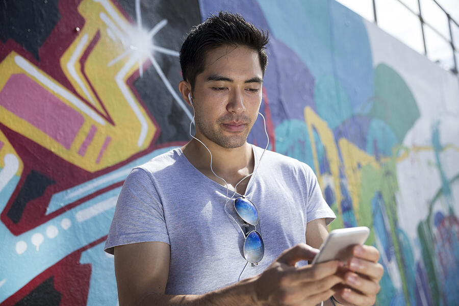 Asian man in front of graffiti reading phone. Photograph by Jenner Images