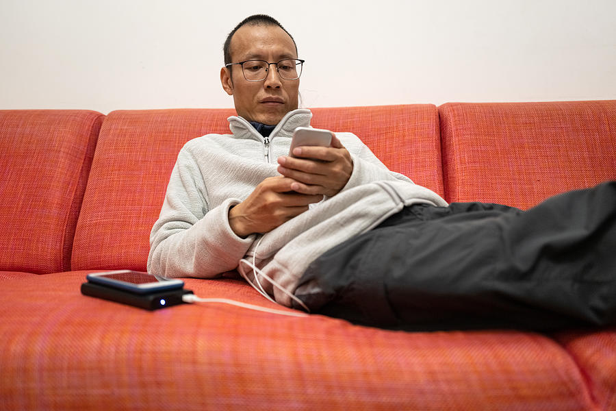 Asian man using charging smartphone on sofa Photograph by Zhihao