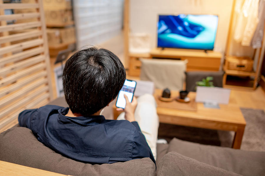 Asian man using smartphone and watching TV at home Photograph by Zephyr18