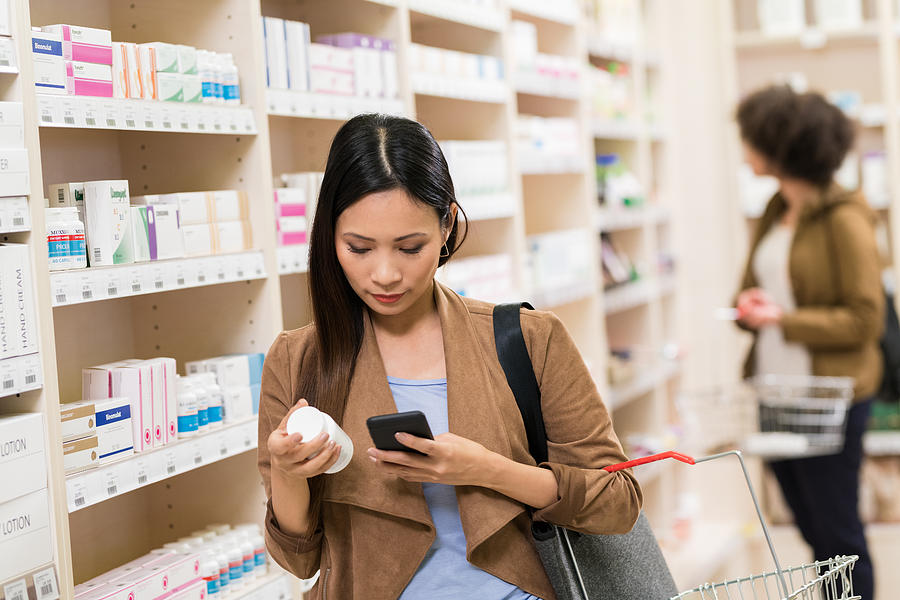Asian Woman Checking Supplement On Smartphone At Pharmacy Photograph by Tashi-Delek