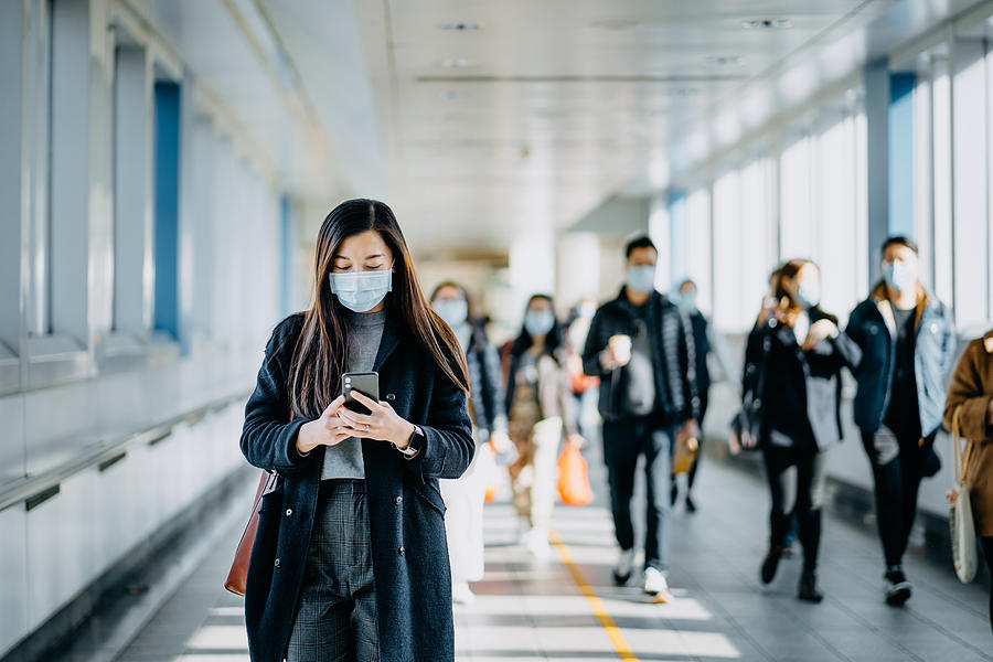 Asian woman with protective face mask using smartphone while commuting in the urban bridge in city against crowd of people Photograph by D3sign