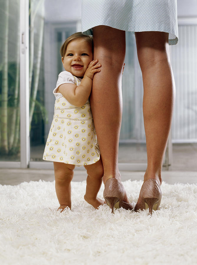 Baby girl (6-9 months) standing on rug, holding onto mothers legs Photograph by Kraig Scarbinsky