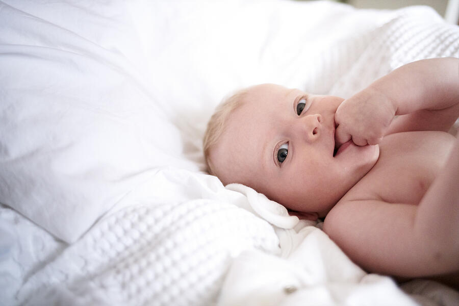 Baby girl lying on parents bed with fingers in mouth looking to camera Photograph by Justin Lambert
