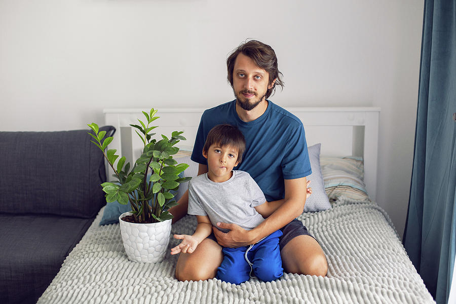 bearded father and son in T-shirt are sitting on a bed with a flower in ...