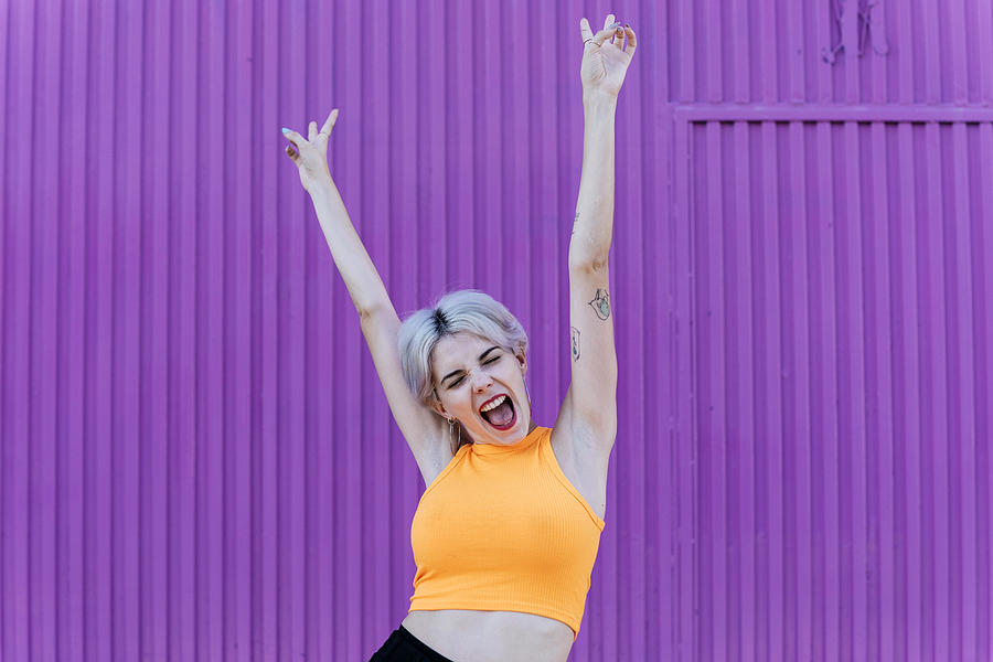 Beautiful blond woman standing in front of purple wall, raising arms, laughing Photograph by Westend61