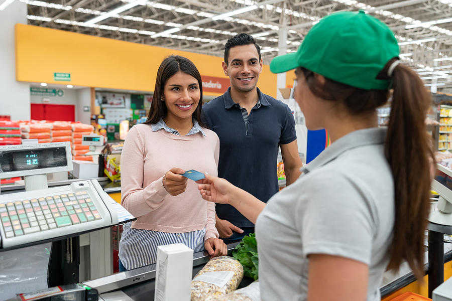 Beautiful couple customer paying for groceries at the cash register with credit card smiling Photograph by Hispanolistic