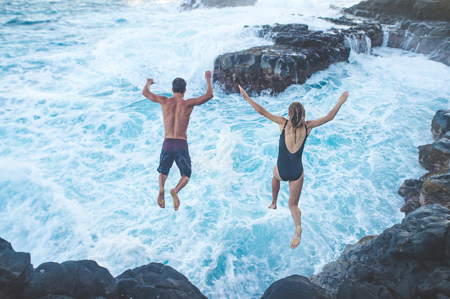 Beautiful couple jumping off cliff into ocean Photograph by FatCamera