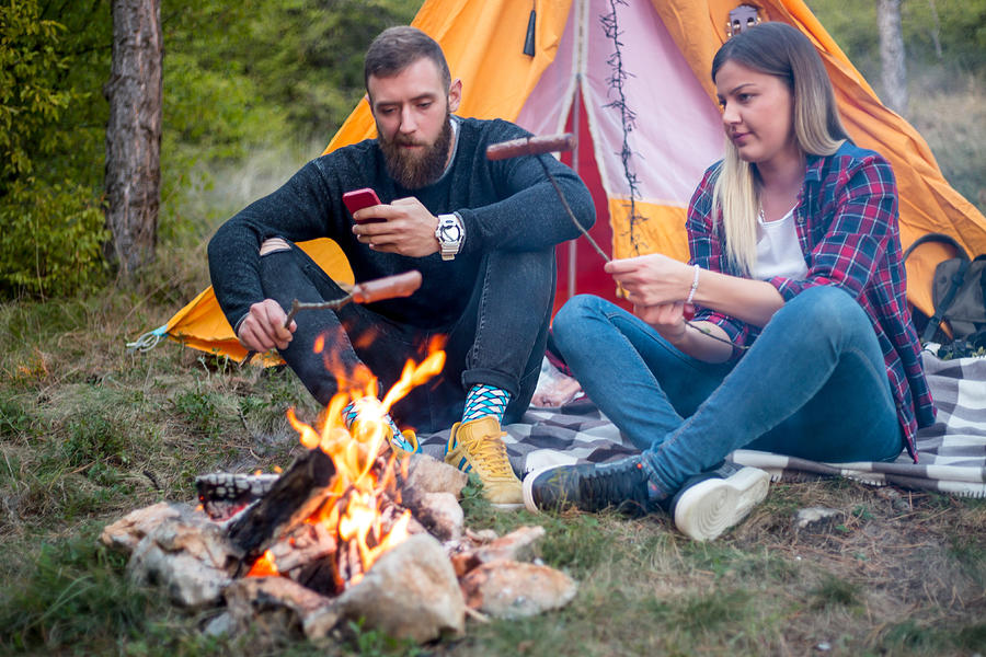 Beautiful couple, sitting, camping around the campfire. On the background with a tent The man flicks the fire with a stick. Photograph by MajaMitrovic