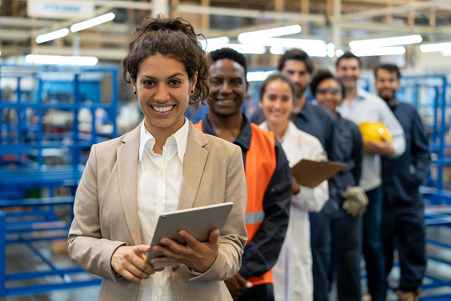 Beautiful female manager at a factory holding a tablet and team of blue collar workers, engineers and inspectors standing in a row smiling at camera Photograph by Hispanolistic