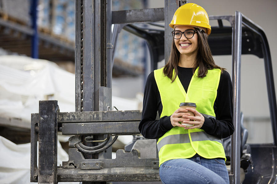 Beautiful girl at factory in a vest and helmet near forklift with coffee. Photograph by Milanvirijevic