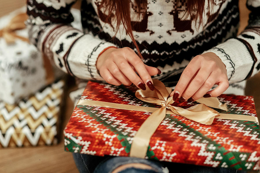 Beautiful girl in reindeer winter sweater unwrapping christmas present with golden ribbon, hands close-up, greeting card concept Photograph by Bogdan Kurylo
