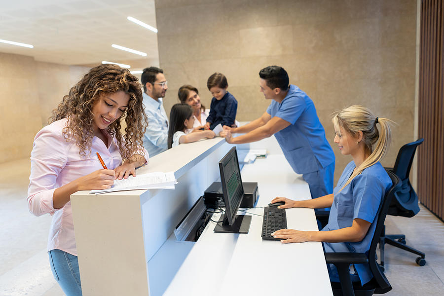 Beautiful latin american patient filling in a form at the hospitals front desk all smiling Photograph by Hispanolistic